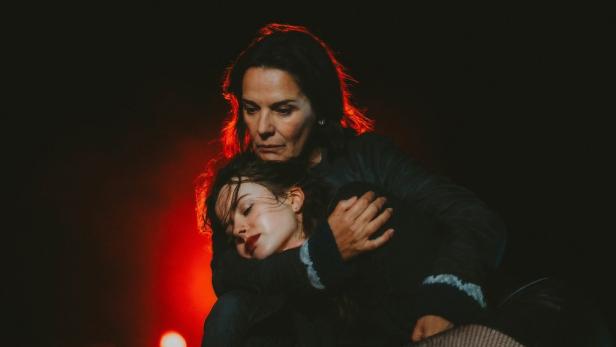 Eine ältere Frau hält eine jüngere Frau im Arm, beide vor dunklem Hintergrund mit rotem Licht.