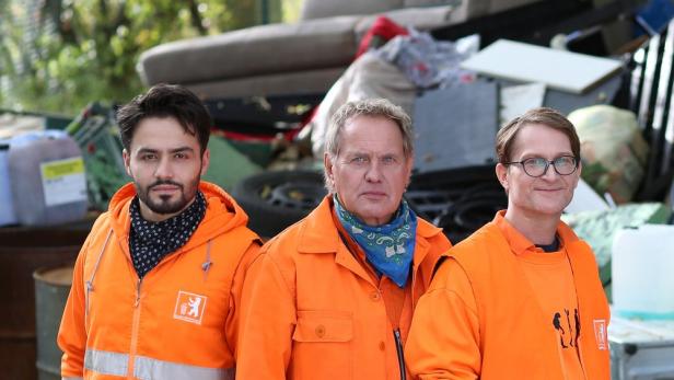 Drei Männer in orangefarbenen Müllabfuhr-Uniformen stehen vor einem Müllhaufen.