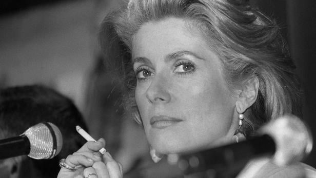 Catherine Deneuve hält eine Zigarette in der Hand.