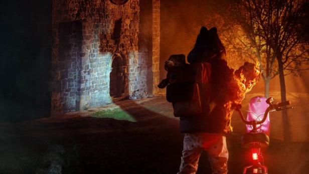 Eine Person mit einem Fahrrad steht vor der Ruine einer alten Kirche bei Nacht.
