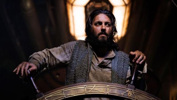 Ein bärtiger Mann in historischer Kleidung steht am Steuerrad eines Schiffes.