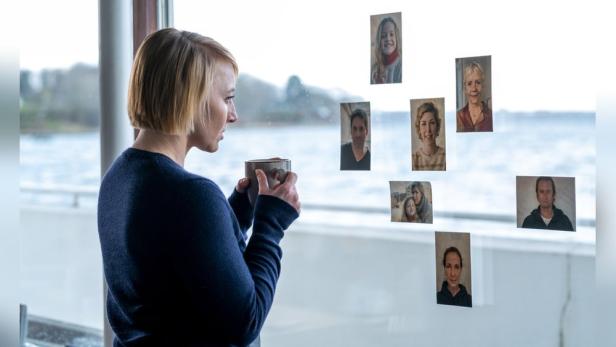 Eine Frau mit einer Tasse in der Hand steht vor einem Fenster mit Fotos.