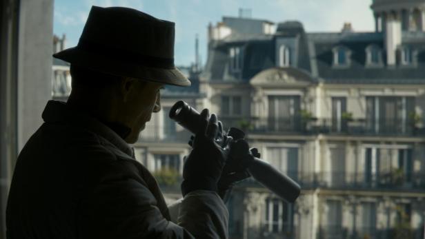Michael Fassbender mit Hut und Fernrohr, der in Paris aus dem Fenster schaut