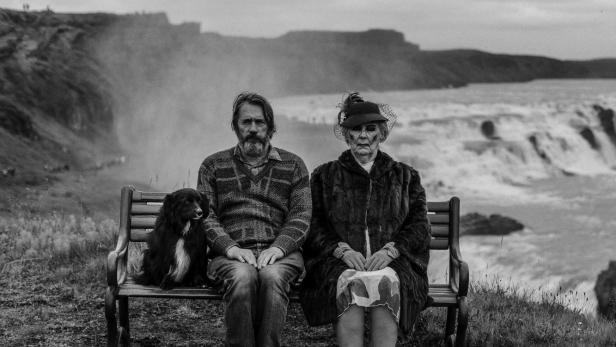 Ein Mann, eine ältere Frau und ein Hund sitzen auf einer Bank vor einem Wasserfall.