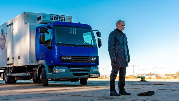Ein Mann steht neben einem blauen Kühlwagen; im Hintergrund eine Baustelle.