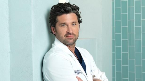 Patrick Dempsey als Dr. Shepherd in einem Arztkittel, an einer Fliesenwand lehnend.