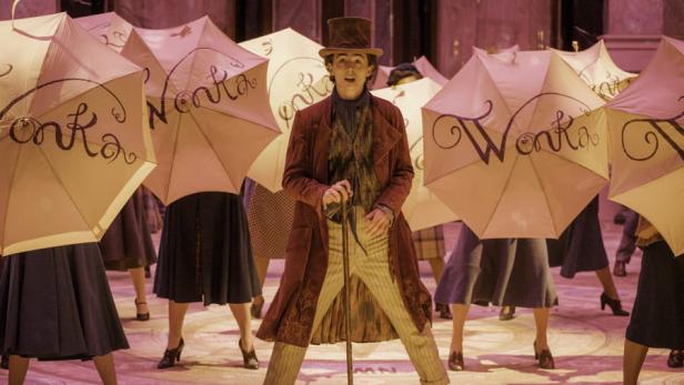 Timothée Chalamet als junger Willy Wonka mit einem lila farbenen Mantel, einem Hut und einem Stock vor Tänzern mit jeweils einem Regenschirm, auf dem "Wonka" drauf steht.