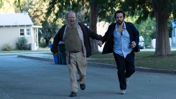 Ein bärtiger Mann und ein Mann mit offenem Hemd rennen Händchen haltend eine Straße entlang.