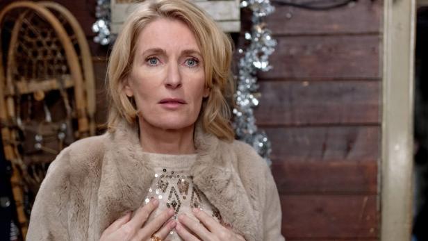 Eine Frau in einem Pelzmantel vor einem weihnachtlich geschmückten Holzhaus.