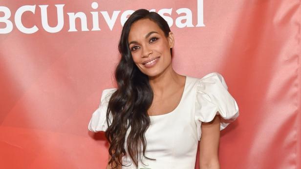 Rosario Dawson lächelt vor einem roten NBCUniversal-Hintergrund.