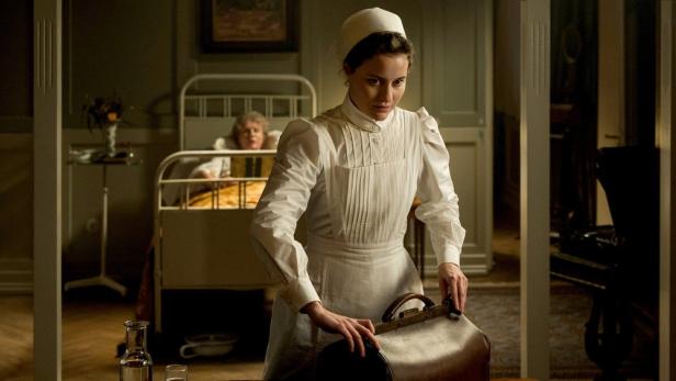 Eine Krankenschwester packt eine Tasche; im Hintergrund liegt eine ältere Frau im Bett.