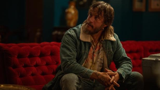 Travis Fimmel sitzt in einer Jeansjacke auf einer roten Couch