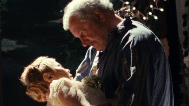 Ein älterer Mann hält ein junges Mädchen im Arm, beide in festlicher Kleidung.