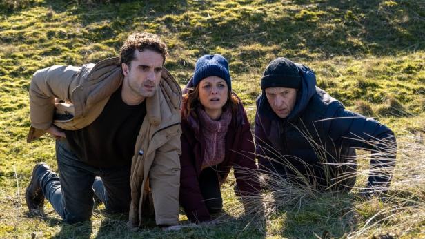 Drei Personen kauern auf einem grasbewachsenen Hügel.