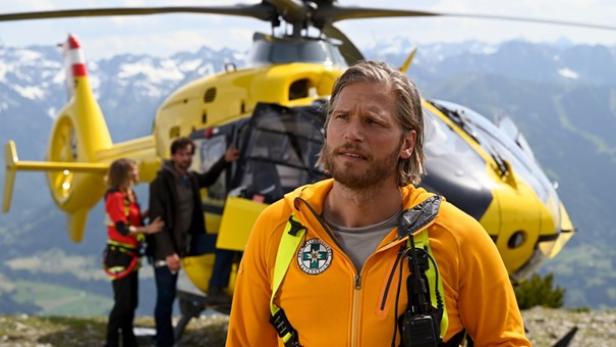 Ein Mann steht in einer orangenen Weste vor einem gelben Rettungshubschrauber in den Bergen.