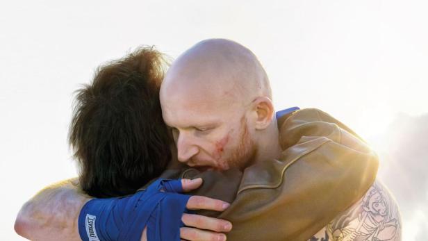 Ein blutiger Boxer mit Tattoos umarmt einen anderen Mann vor dem Filmplakat „Bisons“.