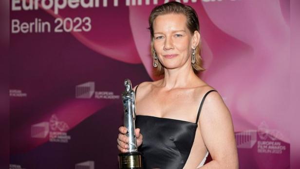 Sandra Hüller in einem schwarzen Kleid, das an der Seite offen ist; vor der pinken Leinwand des Europäischen Filmpreises