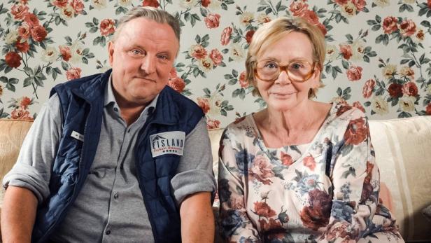 Ein Mann und eine Frau sitzen vor einer floralen Tapete auf einem Sofa.