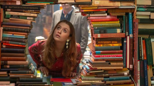 Eine Frau schaut durch eine meterhohe Bücherwand mit einem Loch