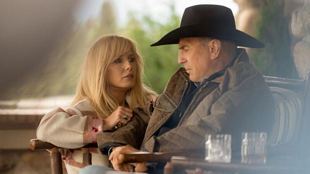 Eine blonde Frau sitzt auf einer Veranda neben einem Mann mit einem Cowboy-Hut. Sie schaut ihn an, er auf den Boden.