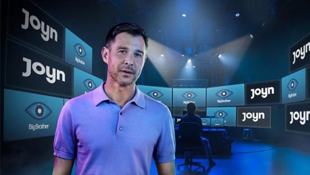 Jochen Schropp steht in einem lila Polohemd vor dutzenden Bildschirmen, auf denen Joyn steht und das Logo von „Big Brother“ zu sehen ist.