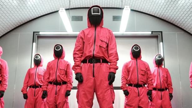 Fünf Personen stehen in V-Stellung und roten Uniformen und schwarzen Gesichtsmasken nebeneinander.
