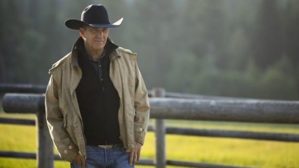 Ein Mann steht vor einem Holzzaun und trägt eine beige Jacke, Jeans, einen Cowboy-Gürtel und Cowboy-Hut