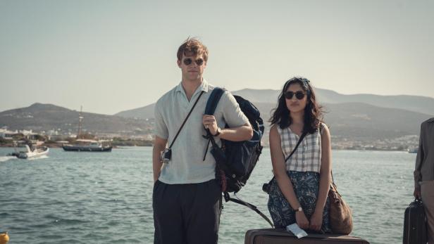 Ein blonder, junger Mann und eine junge Frau mit indischen Wurzeln stehen mit Sonnenbrillen an einem Hafen vor einer griechischen Insel.