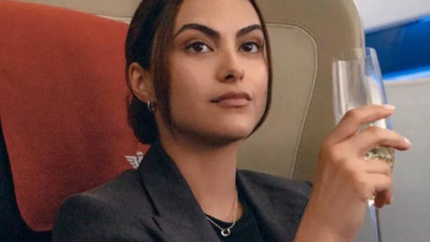 Eine Frau sitzt in einem grauen Blazer in der ersten Klasse in einem Flugzeug und hält ein Champagnerglas in der Hand.