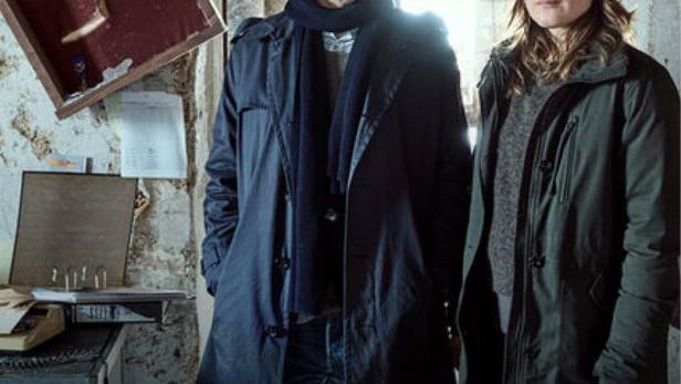 Zwei Kommissare stehen vor dem Poster für „Ein Krimi aus Passau: Gier nach Gold“.