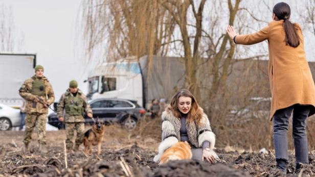 Eine weinende Frau sitzt mit einem Hund auf dem Boden, während eine andere Frau sie beschützt und Soldaten im Hintergrund stehen.