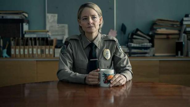 Ein weiblicher Sheriff mit blonden Haaren sitzt mit einem Kaffee in der Hand an einem Schreibtisch.