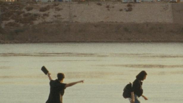 Zwei Personen spazieren an einem Flussufer entlang, eine wirft einen Gegenstand ins Wasser.