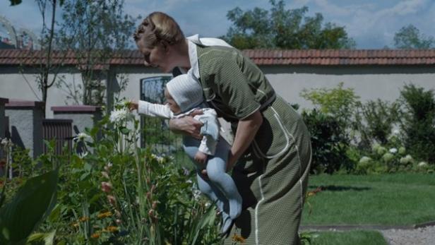 Eine Frau mit Hochsteckfrisur in einem grünen Kleid, die ein Baby in den Armen hält und sich in ihrem Garten über eine Blume beugt.