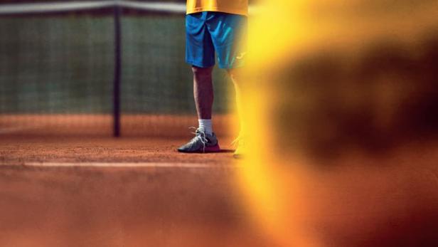 Ein älterer Mann mit Brille jubelt mit einem Tennisschläger auf einem Tennisplatz.