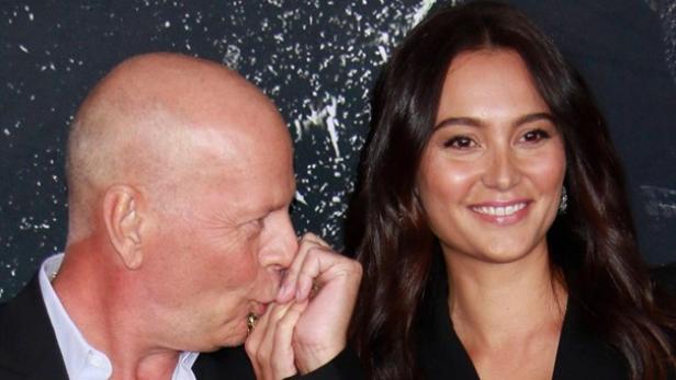Bruce Willis küsst auf einem roten Teppich die Hand seiner Frau, während sie in die Kamera lächelt.