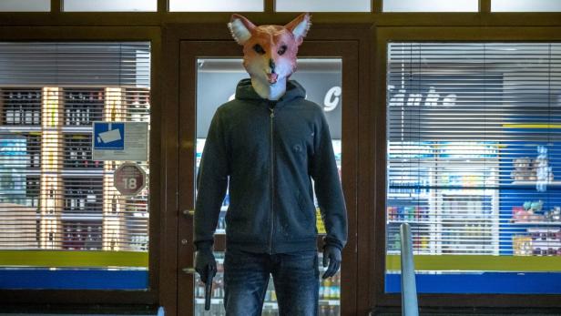 Eine Person mit einer Fuchsmaske und einer Pistole steht vor einem Geschäft.