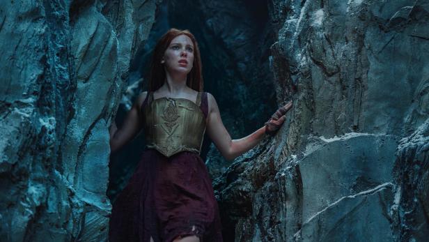 Millie Bobby Brown mit langen, roten Haaren in einem Prinzessinnenkleid, wie sie verängstigt zwischen hohen Felsen steht.