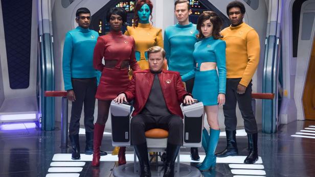 Im Star Trek-Look stehen sechs Personen vor einem Mann auf einem Stuhl. Alle haben bunte, einheitliche Klamotten an und schauen in die Kamera.