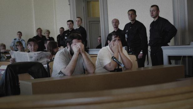 Ein Mann und eine Frau sitzen in einem Gerichtssaal nebeneinander und halten die Hände vor ihr Gesicht. Hinter ihnen stehen ein Dutzend Menschen, das zu ihnen schaut.