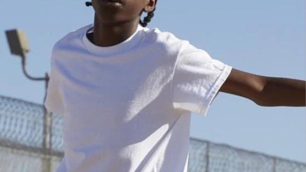 Ein junger Mann mit geflochtenen Zöpfen trägt ein weißes T-Shirt vor einem Zaun.