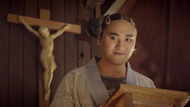 Ein Mann mit Haarklammern steht vor einem Kreuz an einer Wand.