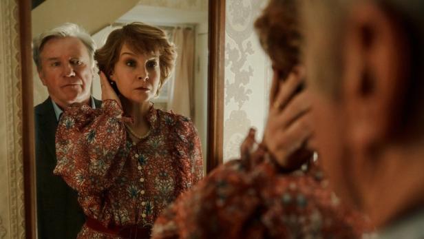 Marianne Voss betrachtet sich im Spiegel, während ein Mann hinter ihr steht.