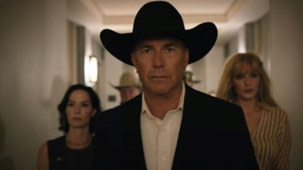 Kevin Costner läuft als Cowboy im Anzug mit Cowboyhut einen Gang entlang. Neben ihm läuft jeweils eine Frau, etwas weiter hinten.