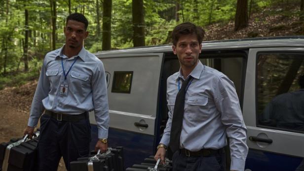 Zwei Wachmänner mit Aktenkoffern stehen vor einem gepanzerten Transporter im Wald.