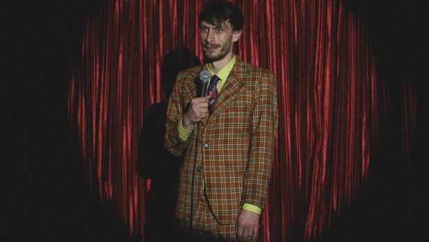 Ein Mann in einem karierten Anzug mit einem Mikro in der Hand auf einer Bühne vor einem roten Vorhang