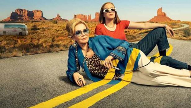 Eine ältere Blondine und eine jüngere, brünette Frau liegen auf einem Highway in den USA. Hinter ihnen braune Steinformationen. Zwei gelbe Streifen der Straße gehen über sie rüber.