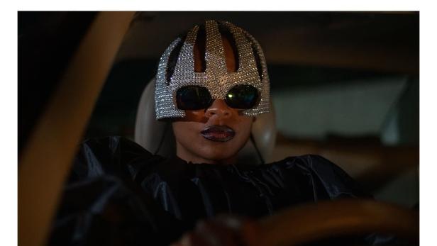 Eine Frau mit einer silbernen, strassbesetzten Maske fährt Auto.