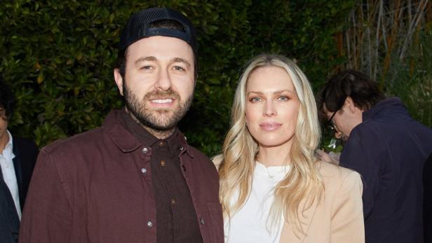 Ein Mann mit Kappe und eine blonde Frau posieren vor einem grünen Hintergrund.