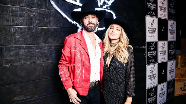 Ryan Bingham und Hassie Harrison posieren vor einer Wand mit Bingham's Bourbon-Werbung.